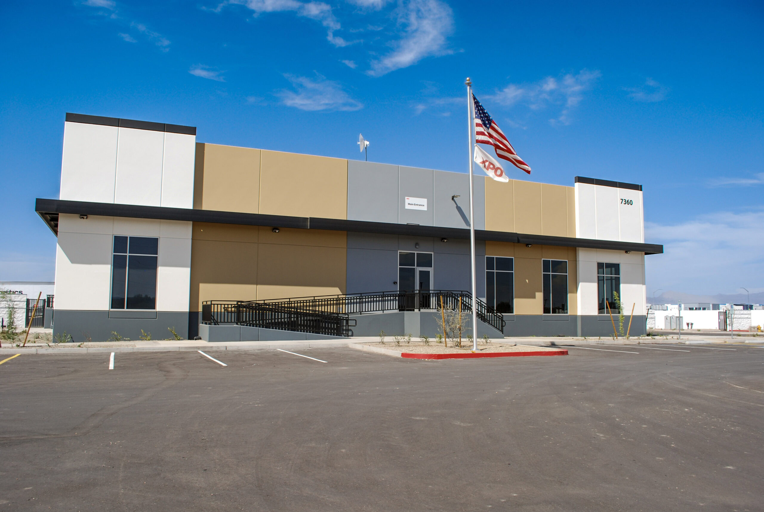 A modern, two-story commercial building with an American flag in front and an empty parking lot under a clear blue sky.