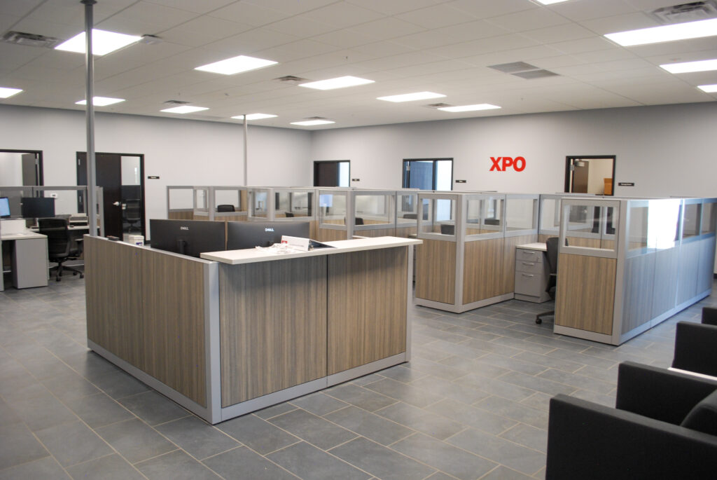 Modern office space with empty cubicles, desks, and chairs; glass partitions and a reception area are visible. “XPO” is displayed in red letters on a wall in the background.