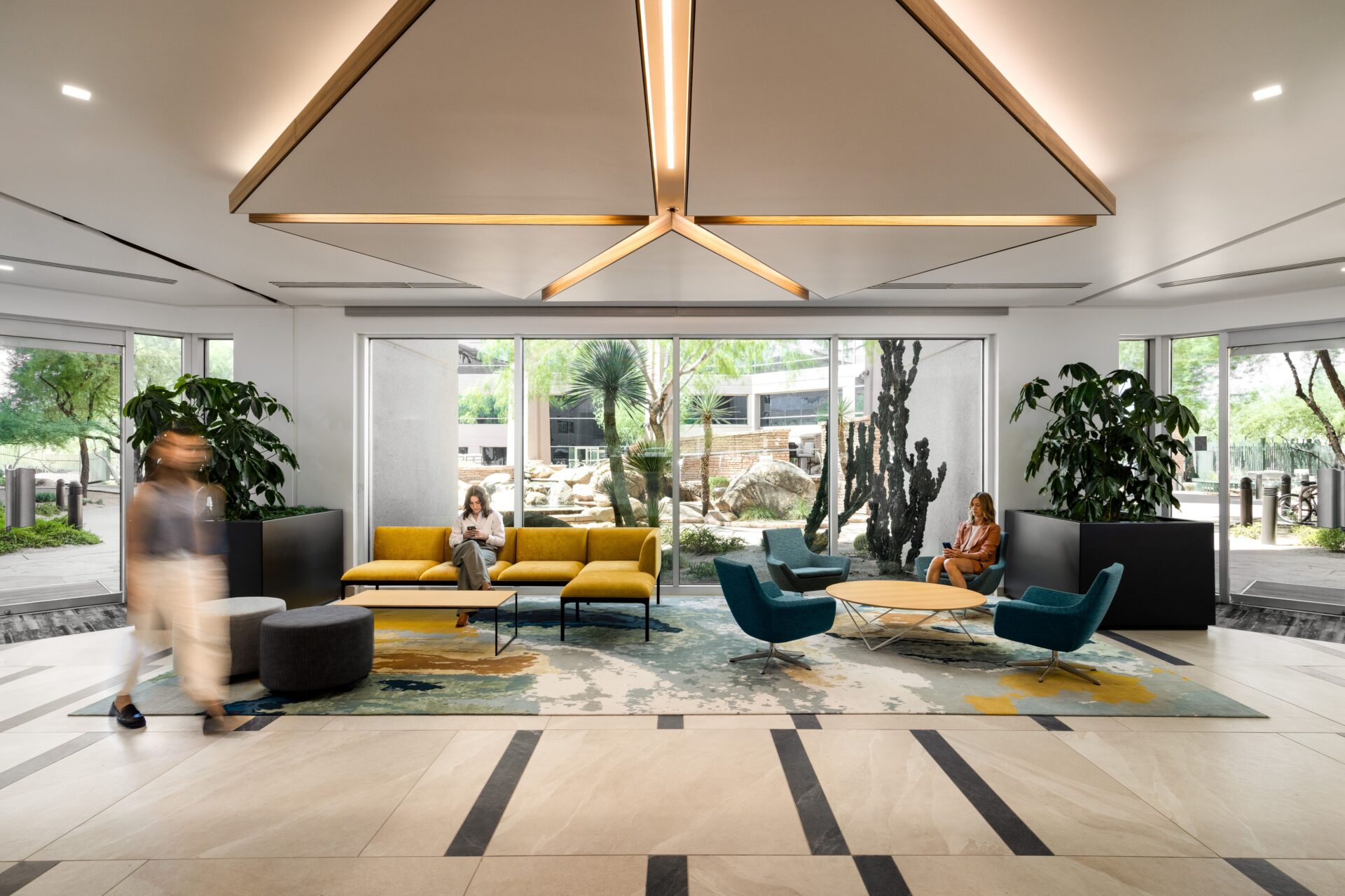 Modern office lobby with geometric lighting, yellow and blue seating, large windows, potted plants, two seated women, and a blurred person walking past.