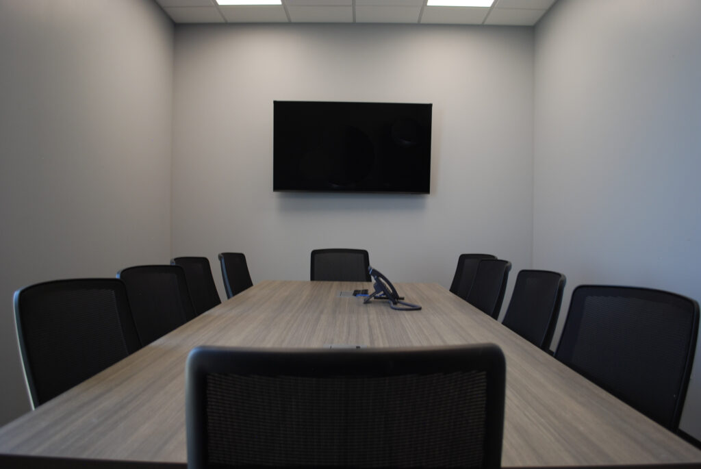 A modern conference room with a long wooden table, eight black chairs, a wall-mounted flat screen, and a phone on the table under bright ceiling lights.