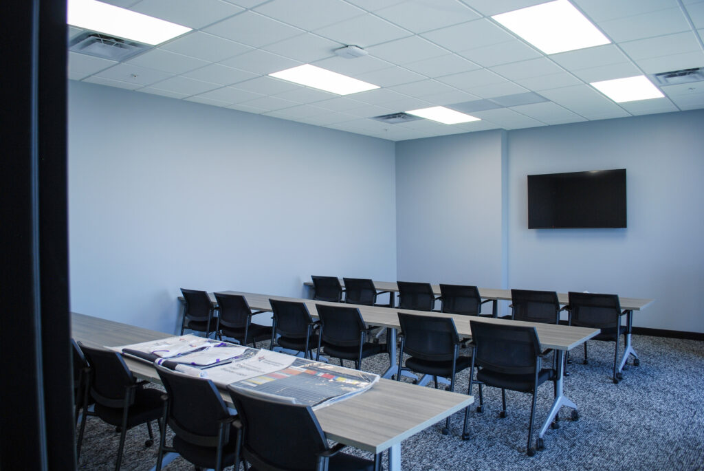 Empty conference room with rows of chairs and tables, a wall-mounted TV, and newspapers laid out on the front table.