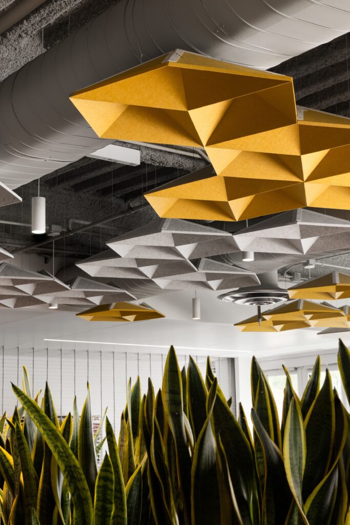 Yellow and gray geometric ceiling panels hang above tall snake plants in a modern office space with exposed pipes and natural light.