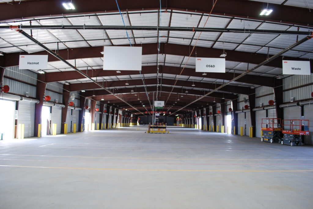 Interior of a large, empty warehouse with high ceilings, overhead signs labeled “Hazmat,” “OS&D,” and “Waste,” open bay doors, and a scissor lift on the right side.