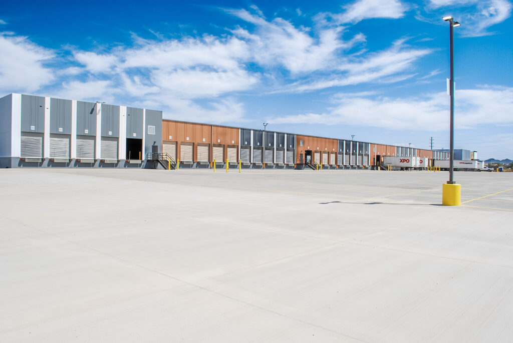 Large, empty industrial loading dock with multiple bay doors, a wide concrete lot, yellow bollards, and a blue sky with scattered clouds overhead.