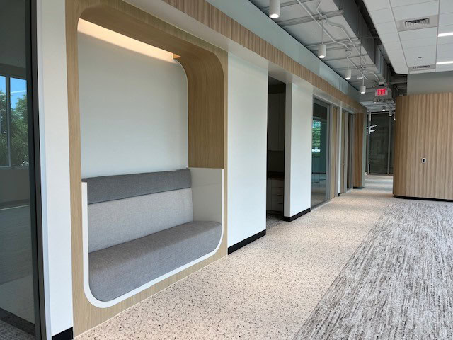 A modern office hallway with a built-in light wood and gray cushioned seating nook, glass-walled offices, and carpeted flooring.