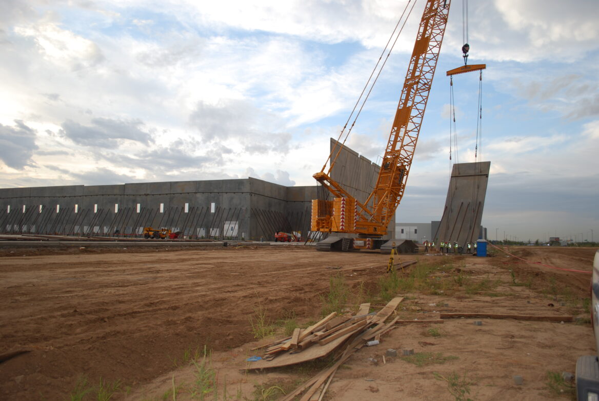 A large yellow crane lifts and positions concrete wall panels at a construction site with workers and equipment visible in the background.
