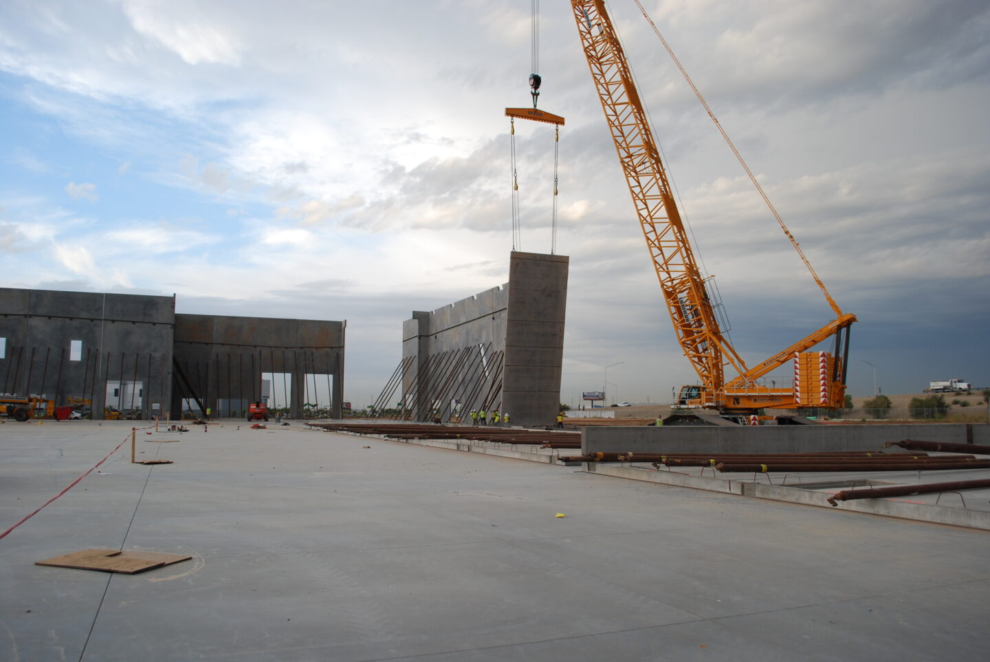 A large yellow crane lifts a concrete wall panel at a construction site with partially erected concrete walls and overcast skies.