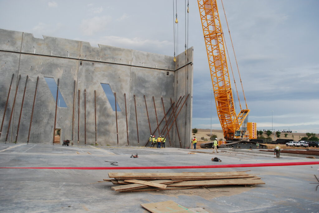 Construction workers and a crane lift a large concrete wall panel at a building site, with steel supports and scattered materials visible on the ground.