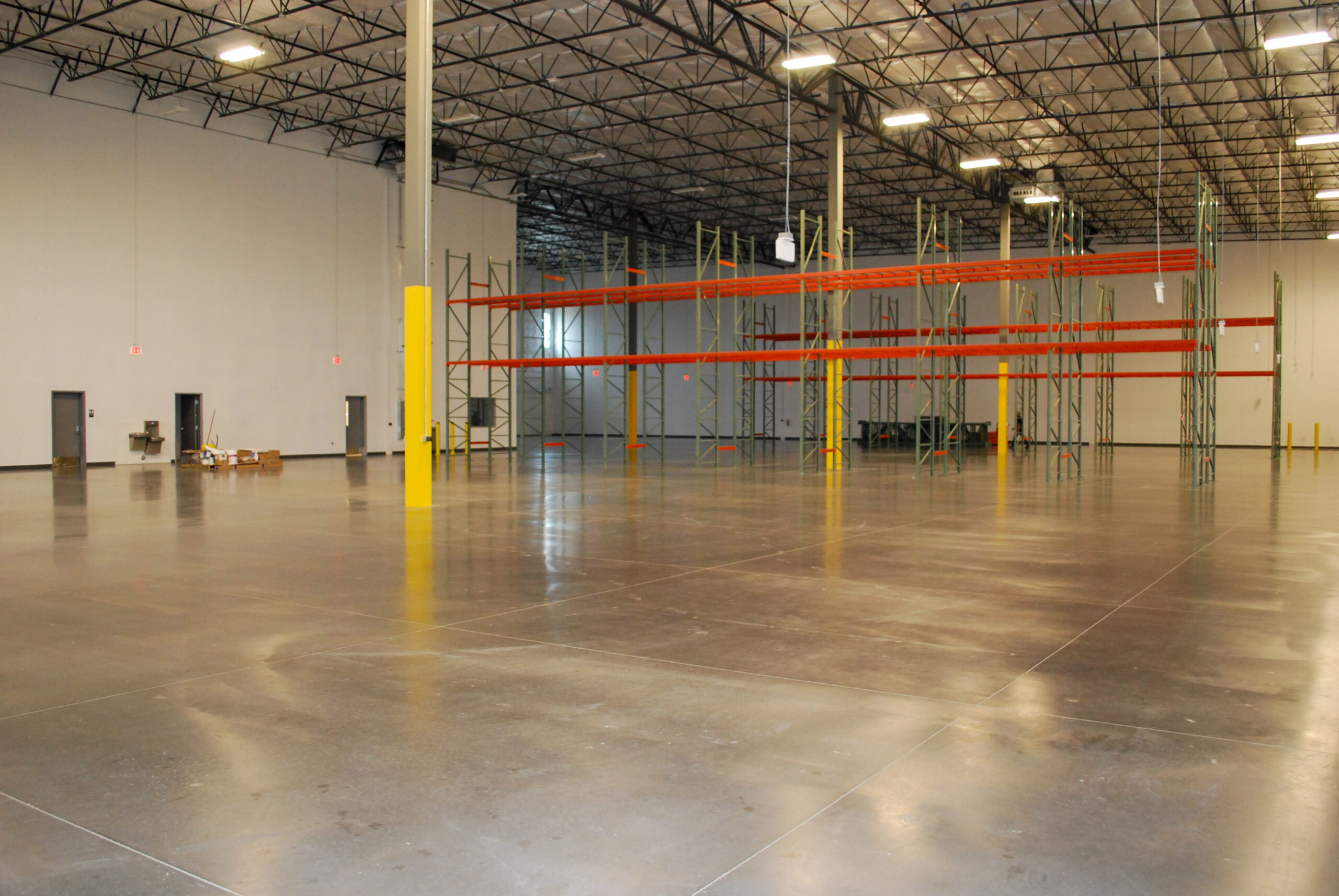 Interior of a large, empty warehouse with high ceilings, exposed beams, and a few metal storage racks along one wall. The concrete floor is clean and shiny.