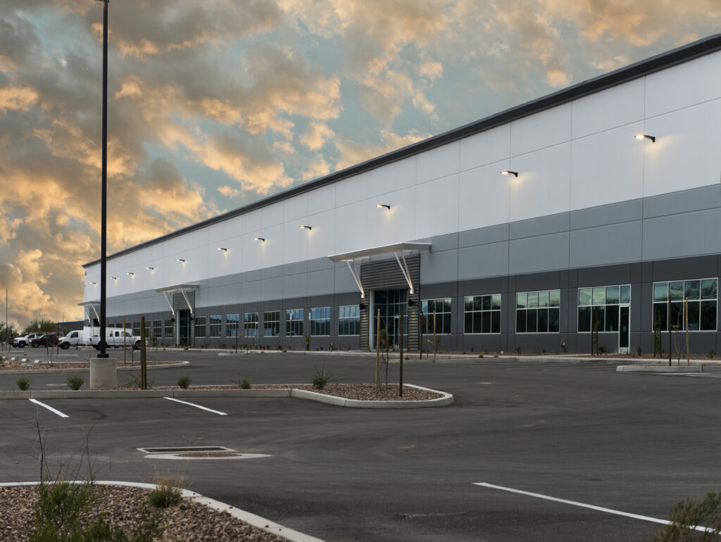 Large, modern industrial warehouse building with gray and white exterior, many windows, and an empty parking lot at sunset.