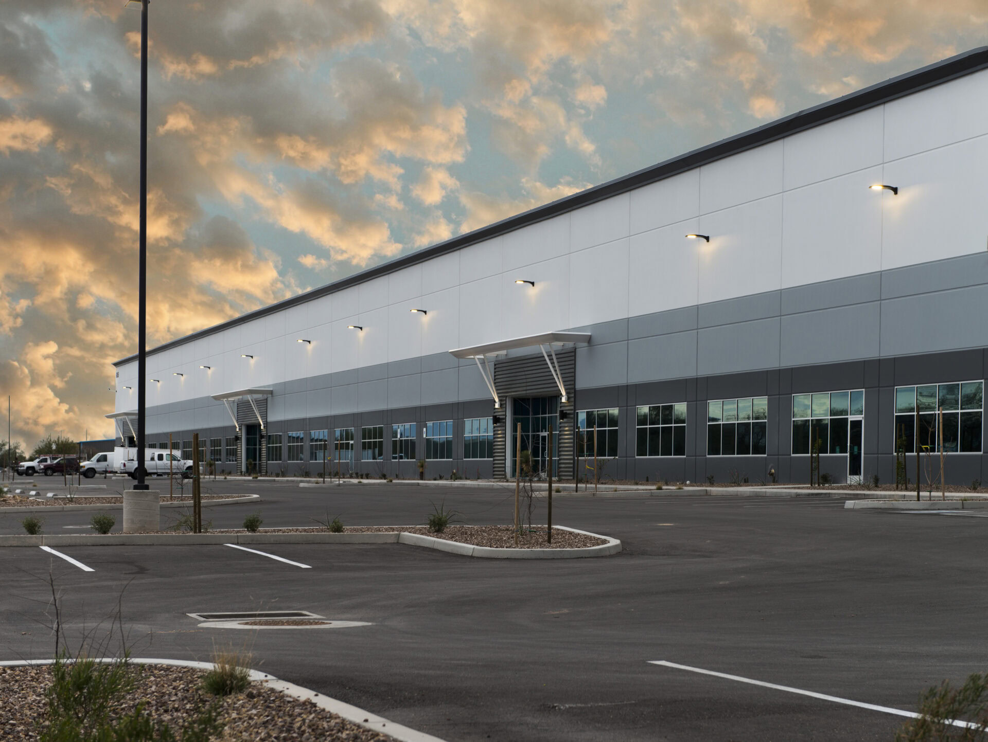 Large, modern industrial warehouse building with gray and white exterior, many windows, and an empty parking lot at sunset.
