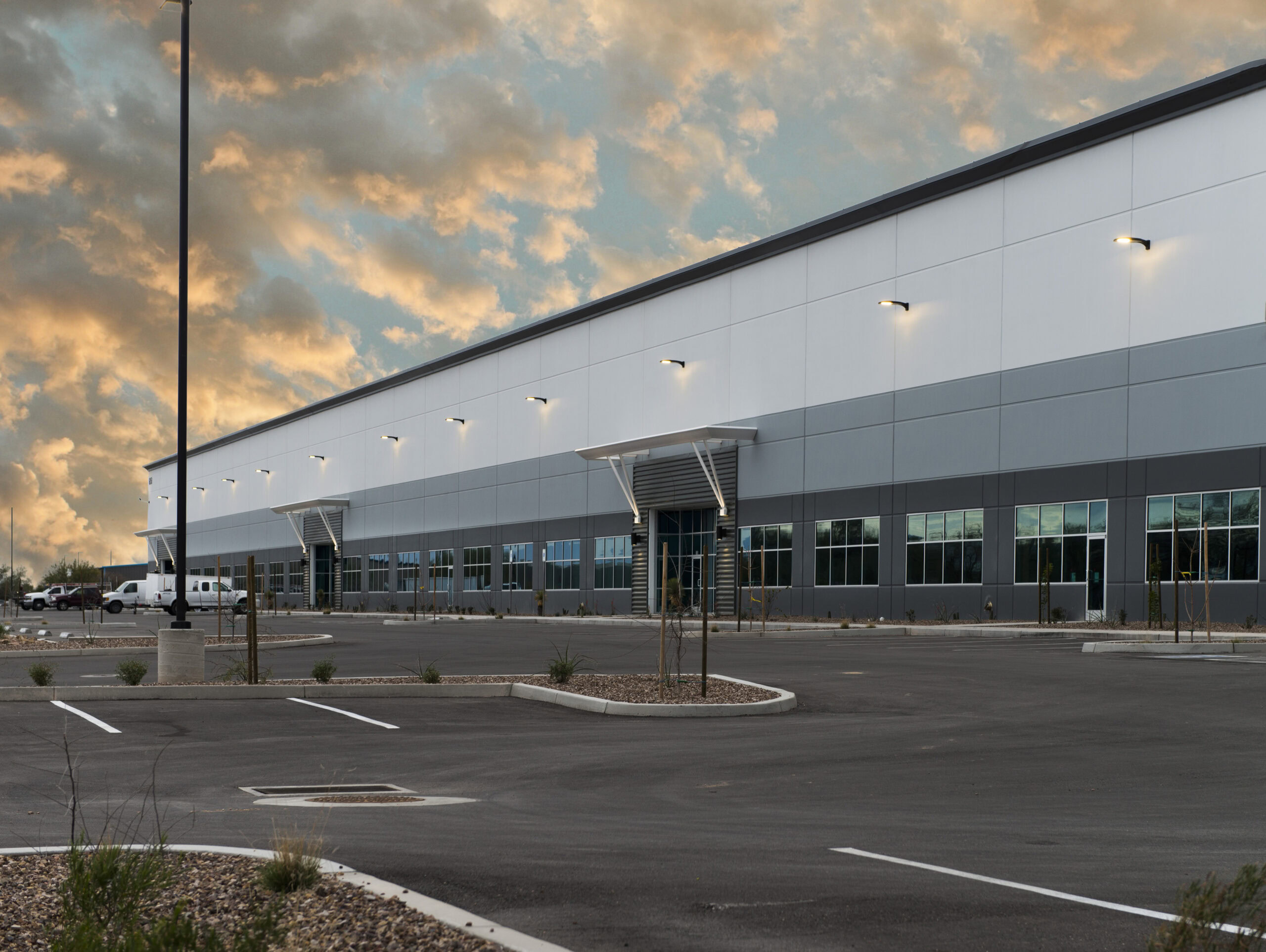 Large, modern industrial warehouse building with gray and white exterior, many windows, and an empty parking lot at sunset.