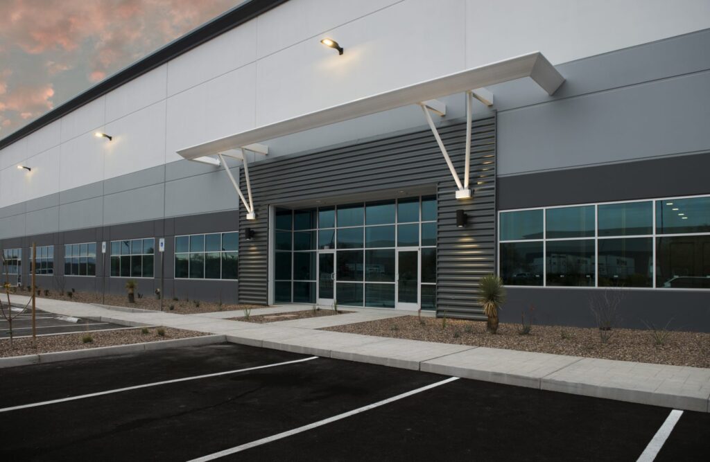 Modern industrial building exterior with large windows, gray and white facade, metal awning, and an empty parking lot in front.