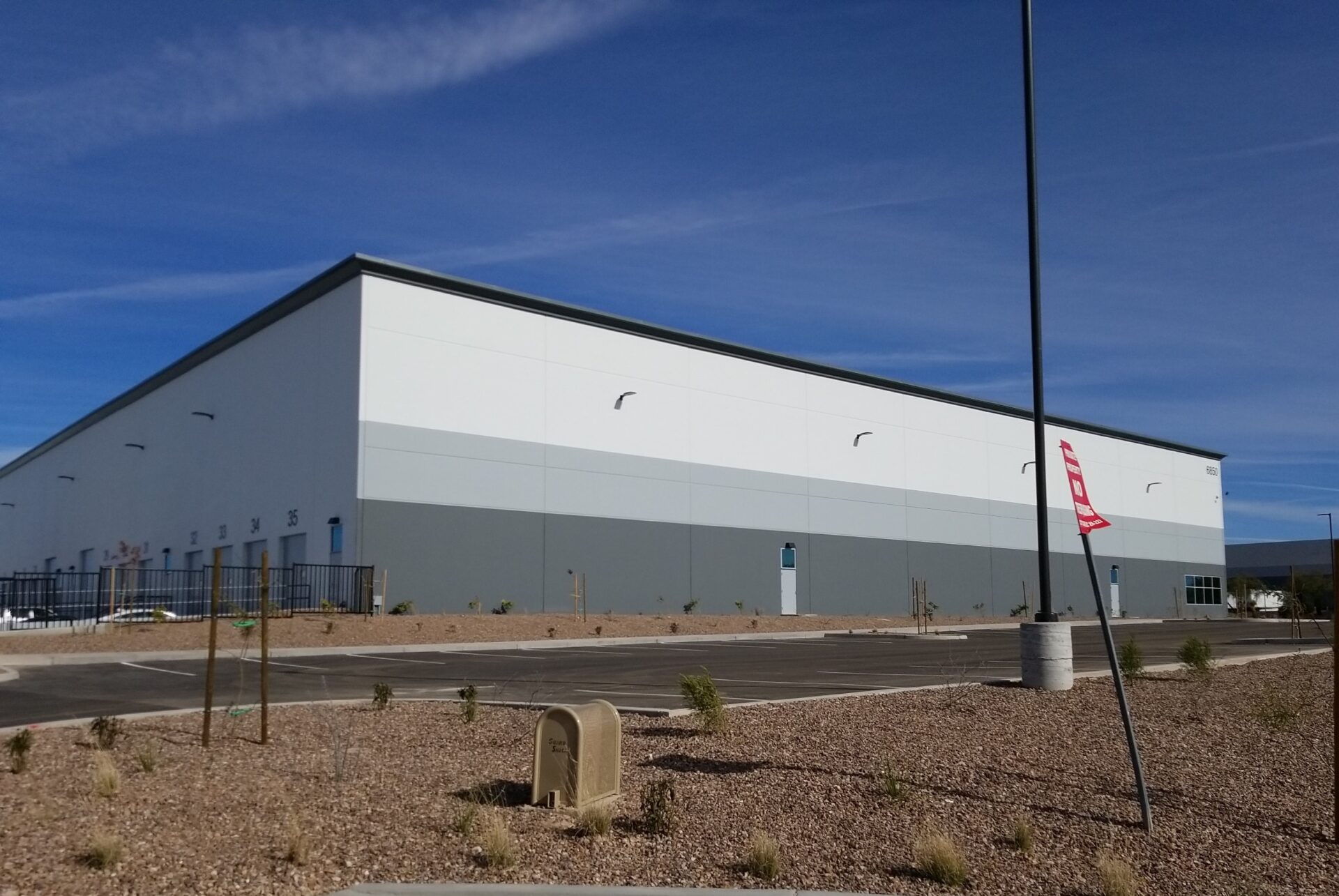 A large, windowless industrial building with a gray and white exterior stands in a dry, landscaped parking lot under a blue sky.