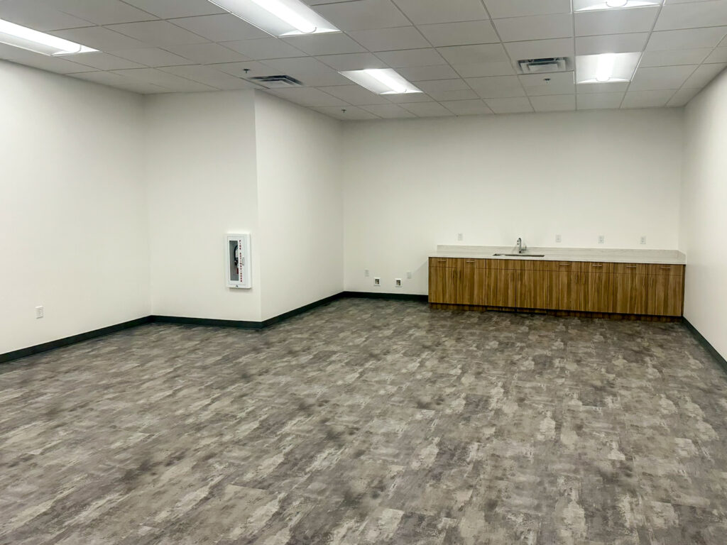 Empty room with gray flooring, white walls, ceiling lights, a wall-mounted fire extinguisher cabinet, and a wooden counter with a sink along the back wall.