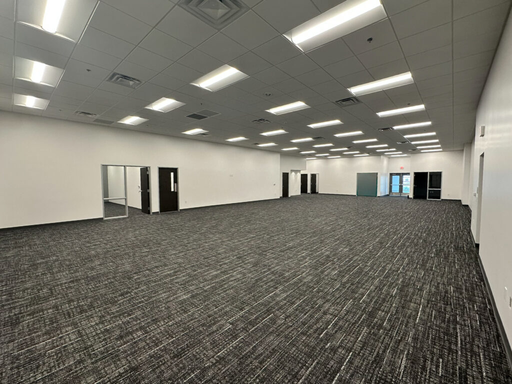Large, empty office space with gray carpet, white walls, fluorescent ceiling lights, and several doors along the far wall. Natural light enters through glass doors at the back.