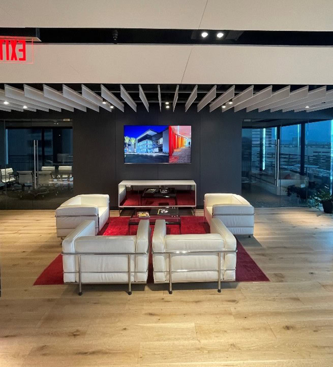 A modern office lounge with four white armchairs and a glass table on a red rug, facing a wall-mounted TV, with glass-walled rooms and city views in the background.