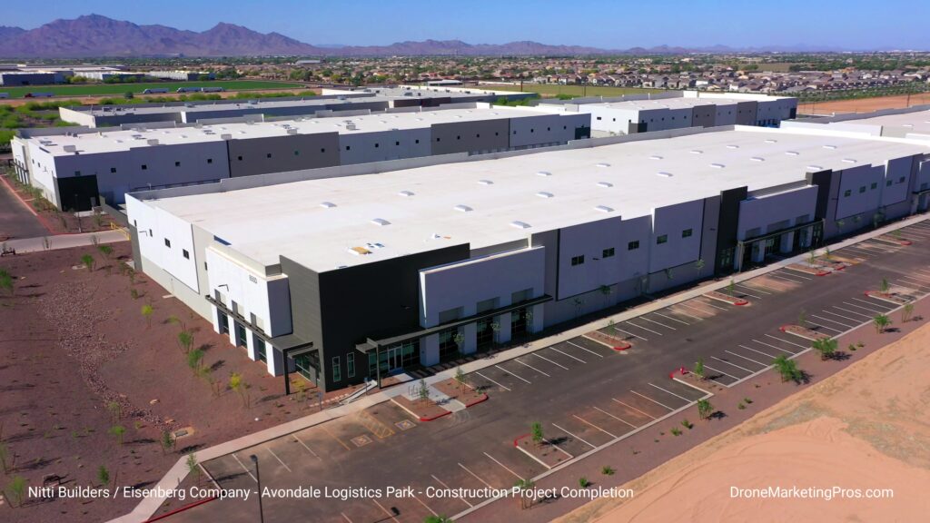 A large, newly constructed warehouse or logistics facility with a white roof and an adjacent empty parking lot, set in a desert landscape with mountains in the background.