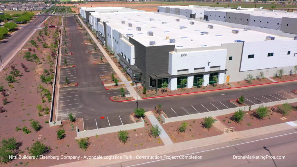 Aerial view of a large white warehouse building with adjacent parking lot, landscaped with small trees and shrubs, in an industrial area.