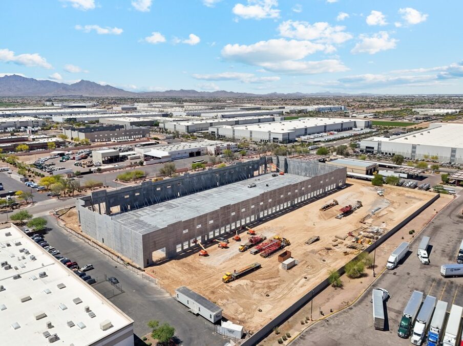 Aerial view of a large industrial building under construction in a commercial area, surrounded by warehouses, trucks, and desert landscape in the background.