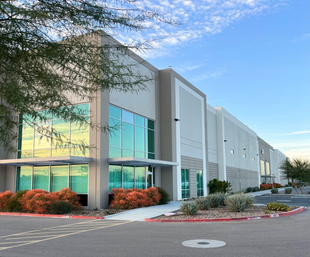 Modern industrial building with large glass windows, gray facade, and landscaped bushes in front, under a clear blue sky.