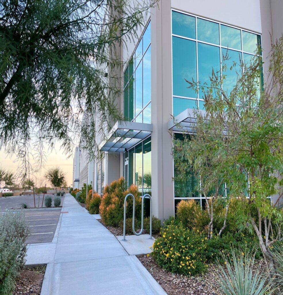 A modern office building with large reflective windows, a glass entry canopy, landscaped plants along the sidewalk, and adjacent parking spaces.