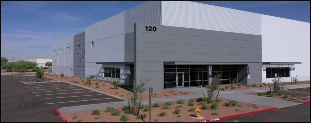 A modern, gray industrial building labeled "120" with large windows, surrounding landscaping, and an empty parking lot under a clear sky.