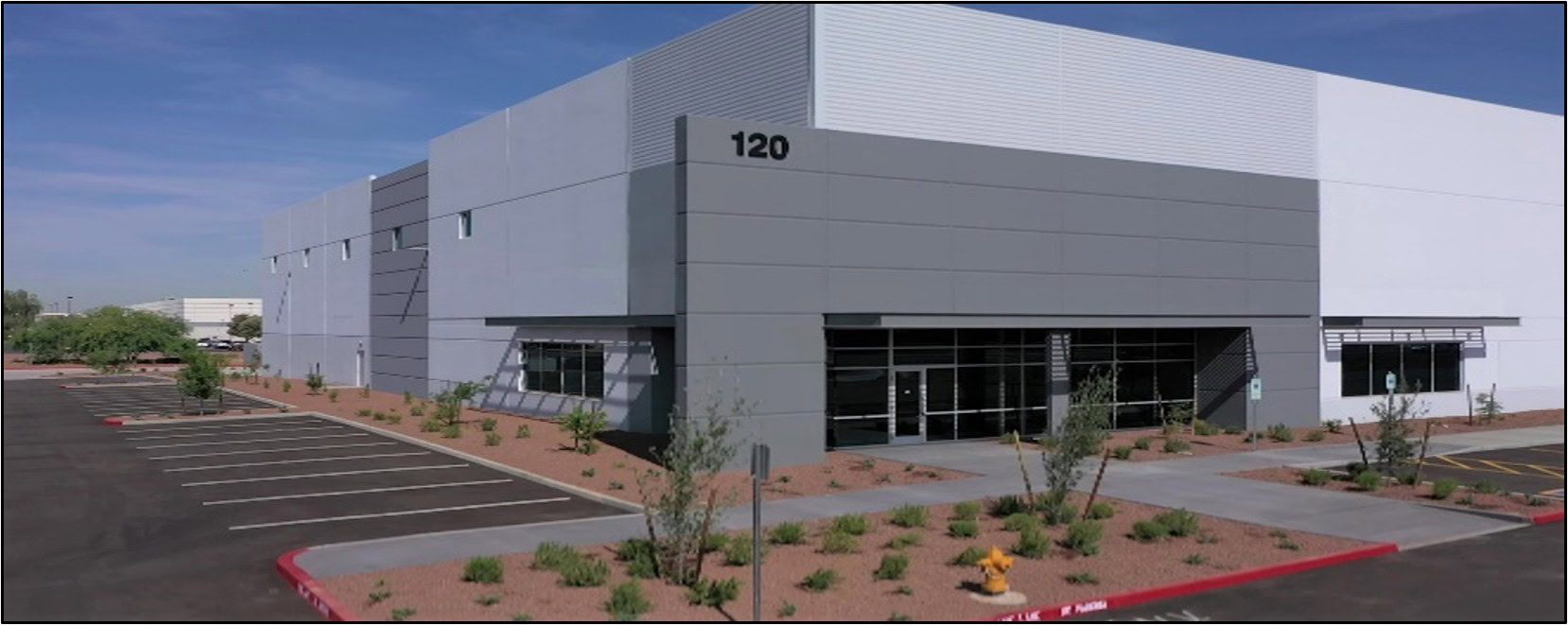 A modern, gray industrial building labeled "120" with large windows, surrounding landscaping, and an empty parking lot under a clear sky.