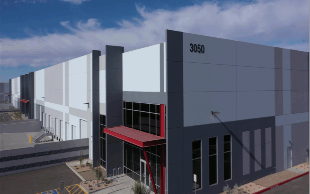 A large modern industrial warehouse building with a red awning, labeled 3050, under a blue sky with some clouds.