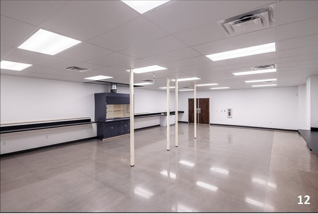 A clean, empty room with polished concrete floors, white walls, support poles, overhead fluorescent lights, and black countertops along the walls. The number 12 appears in the bottom right corner.