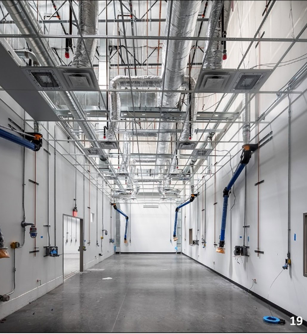 Large industrial room with exposed ductwork and piping on the ceiling, white walls, concrete floor, and several blue retractable hoses mounted along the walls.
