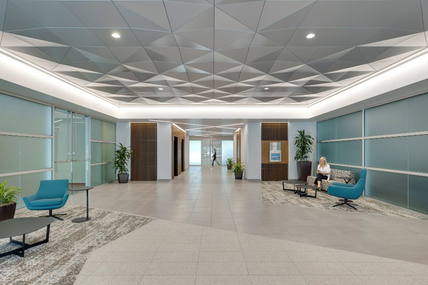 Modern office lobby with geometric ceiling, seating area with blue chairs, glass walls, large plants, and a person sitting on the right side.