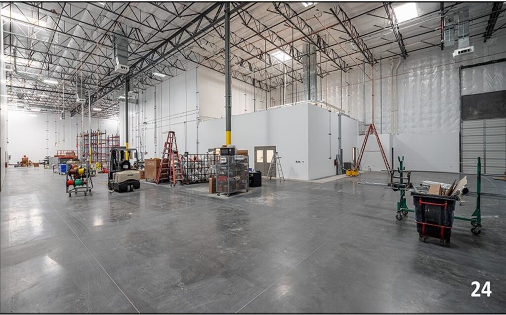 Large warehouse interior with high ceilings, exposed piping, shelves, ladders, carts, and boxes; a small enclosed room is visible in the center right.