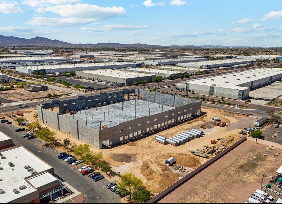 Aerial view of a large warehouse under construction in an industrial area, with surrounding warehouses, parked vehicles, and mountains in the background.