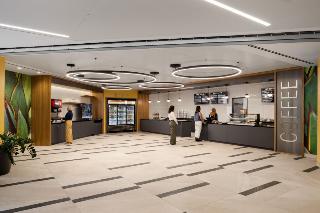 A modern coffee shop with three customers and one staff member at the counter, bright lighting, large menu boards, and a beverage cooler along the back wall.