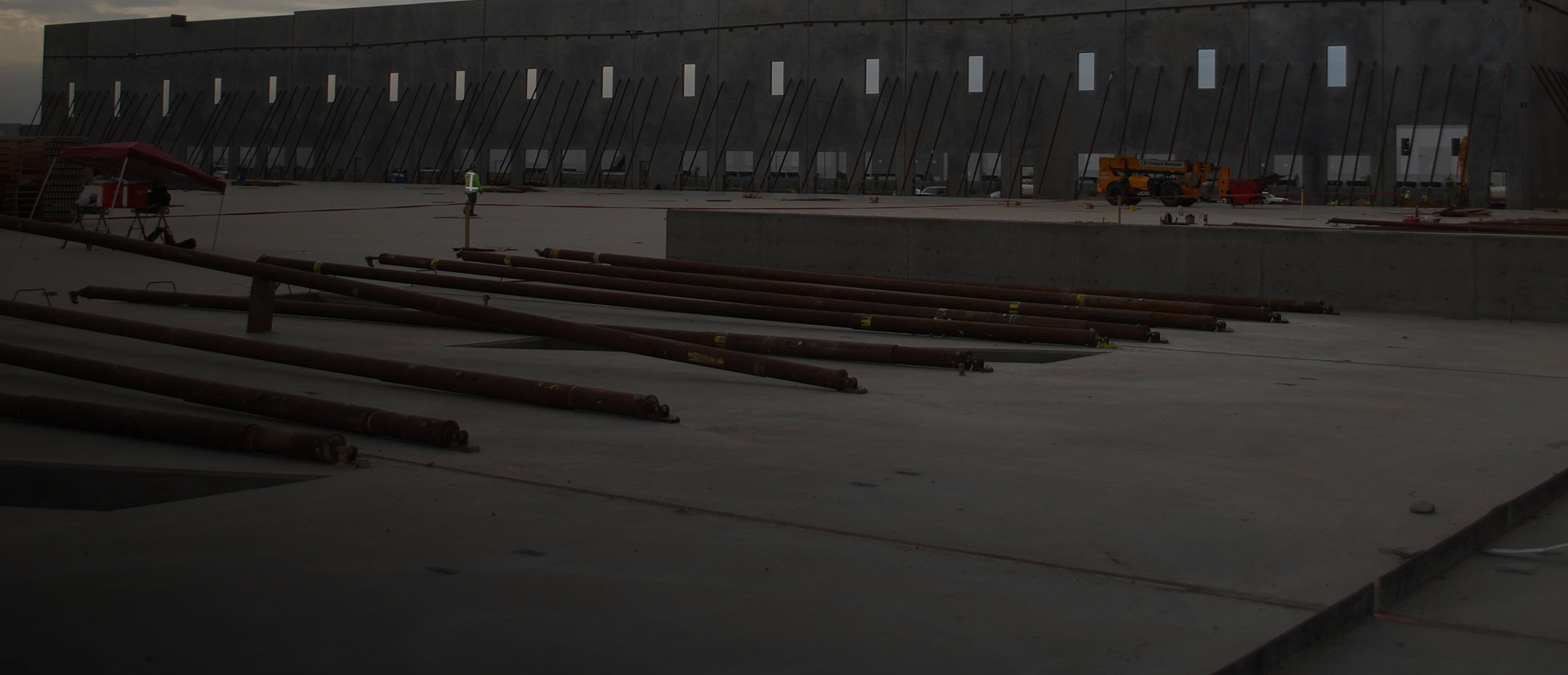A construction site with large concrete walls, steel beams lying on the ground, and workers near a yellow vehicle in the background.