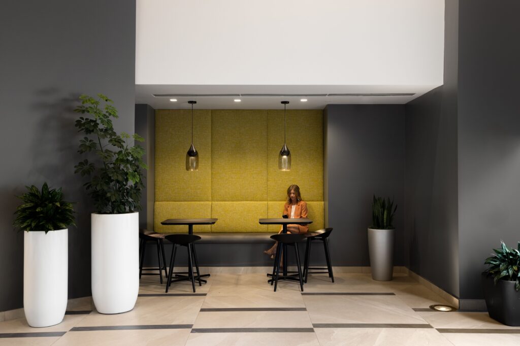 A woman sits alone at a table in a modern lounge area with yellow cushioned walls, pendant lights, and large potted plants.