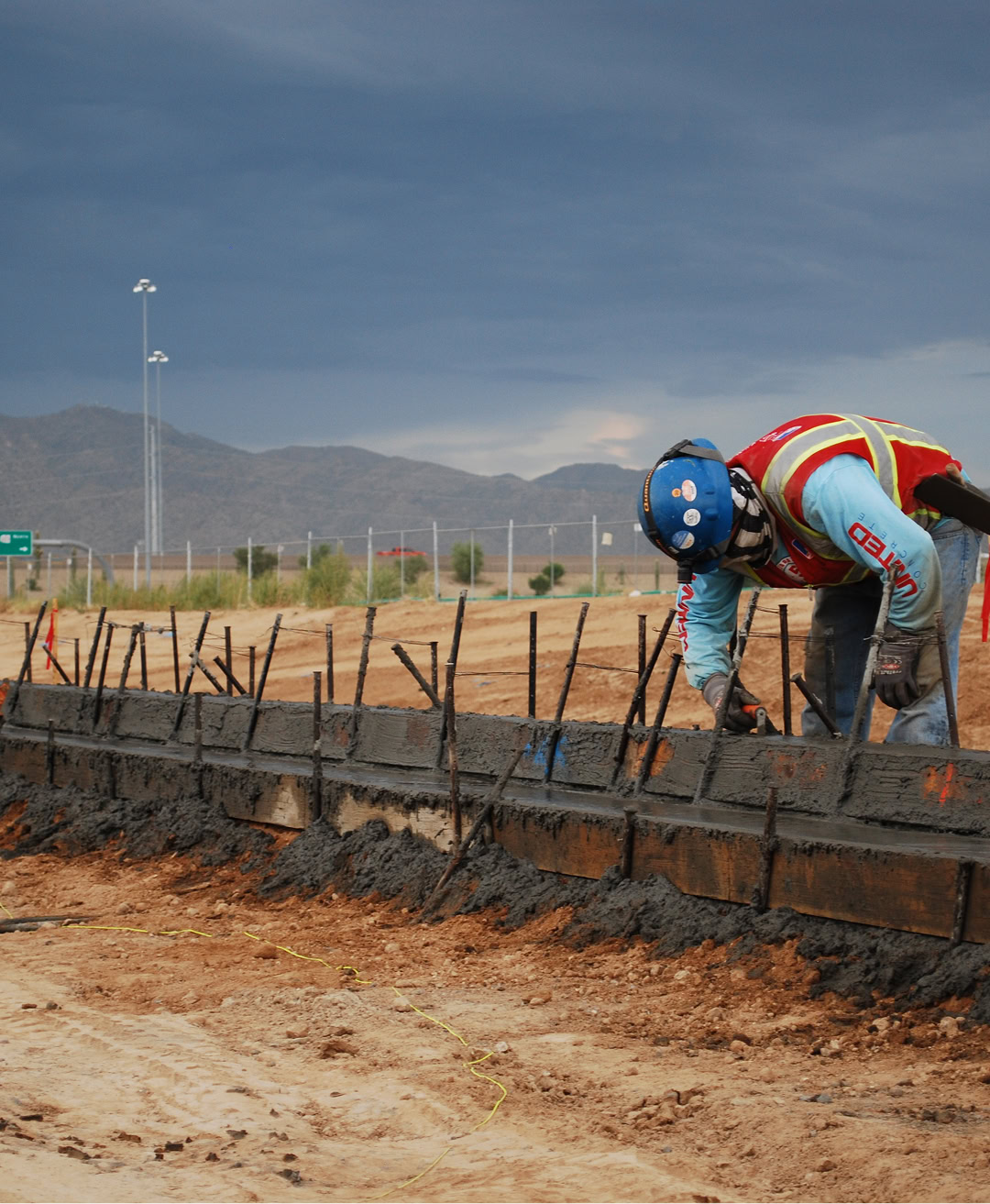 A construction worker in safety gear applies concrete to a curb frame at a roadside construction site, with mountains and dark clouds in the background.