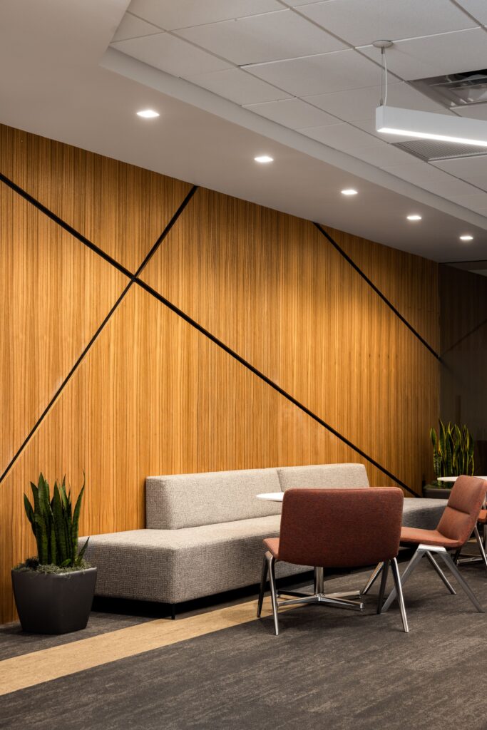 Modern office lounge area with a wooden accent wall, a gray upholstered bench, two red chairs, small tables, potted plants, and ceiling lights.
