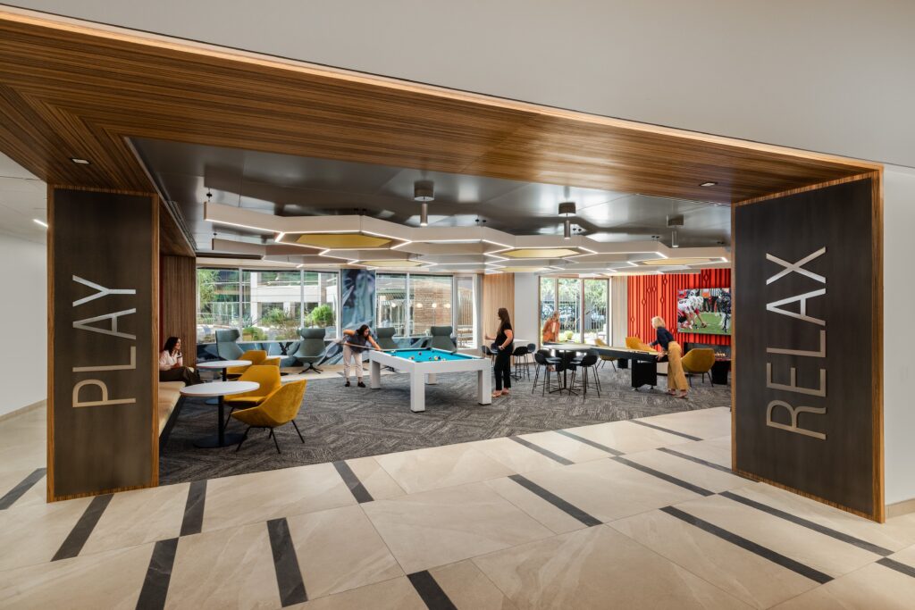 Modern recreation room with people playing pool, sitting at tables, and socializing; signs on walls read "PLAY" and "RELAX"; large windows provide natural light.