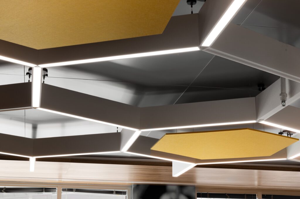 Geometric ceiling lights and yellow acoustic panels are installed in a modern interior space with a metallic and wood finish.