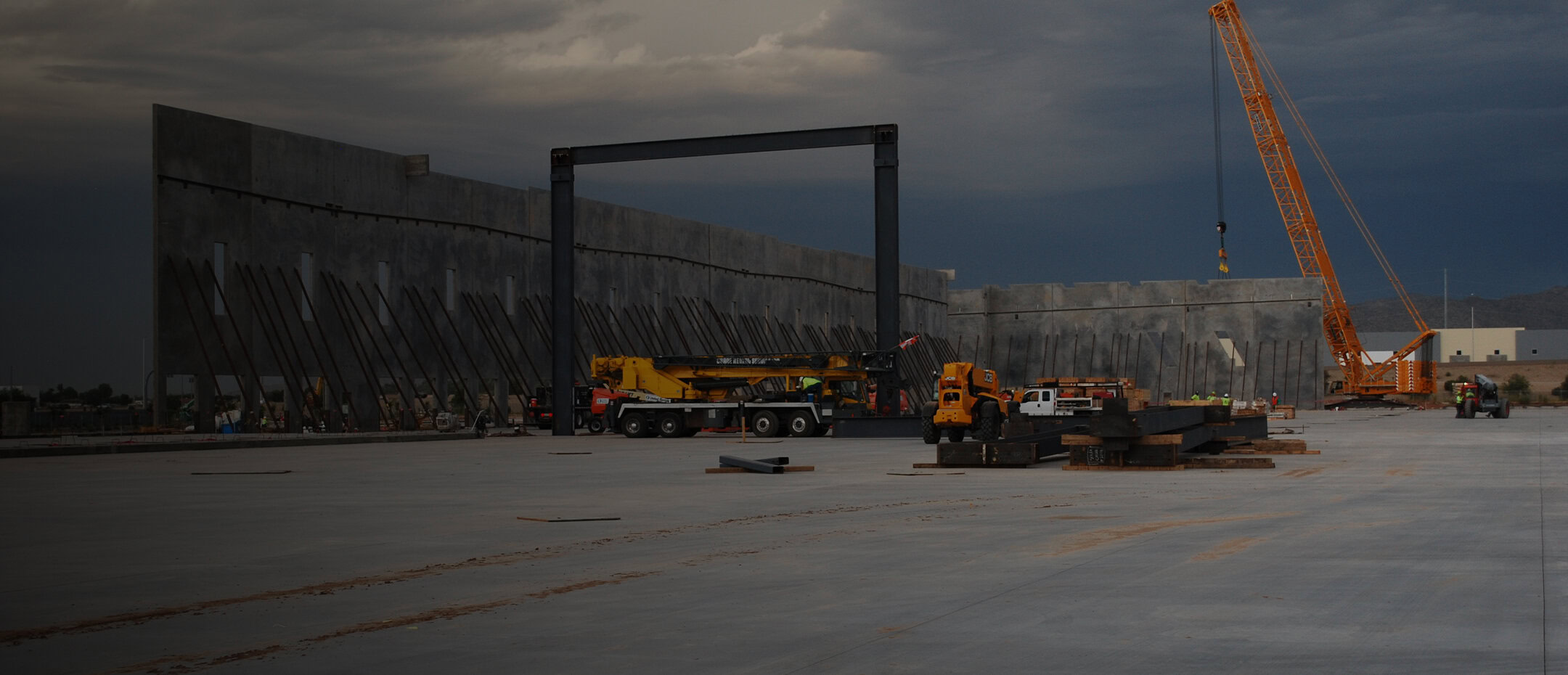 Large construction site with concrete slab, several cranes, heavy machinery, and partially erected concrete walls under a cloudy sky.