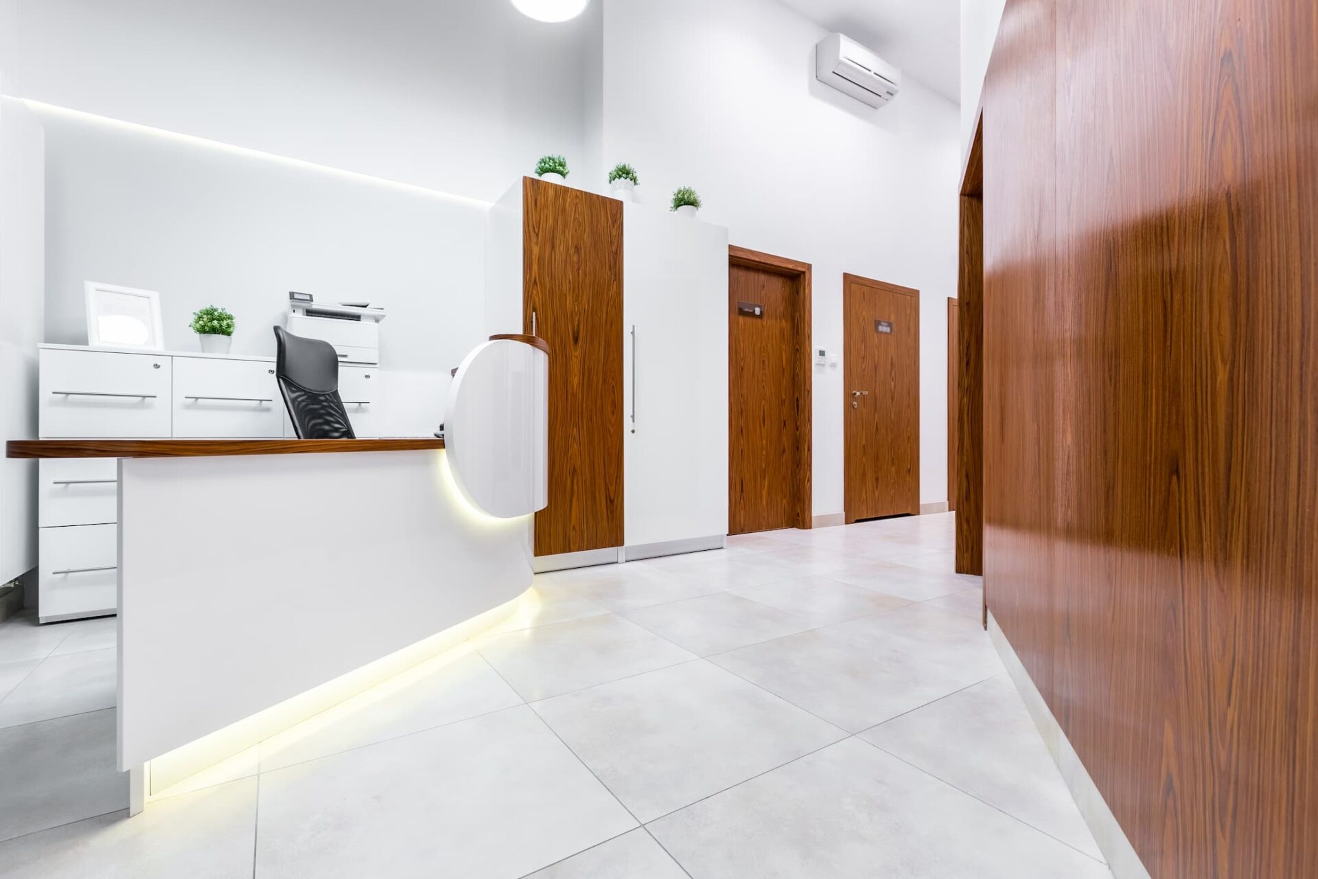 Modern, minimalist office reception area with a white desk, wooden cabinets, potted plants, and several closed wooden doors along a tiled hallway.